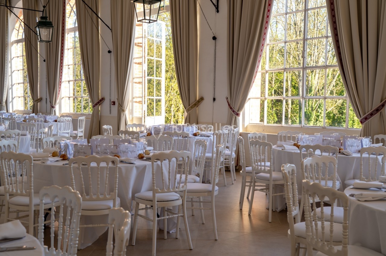 Salle de réception lumineuse avec des tables rondes dressées de nappes blanches et des chaises élégantes.