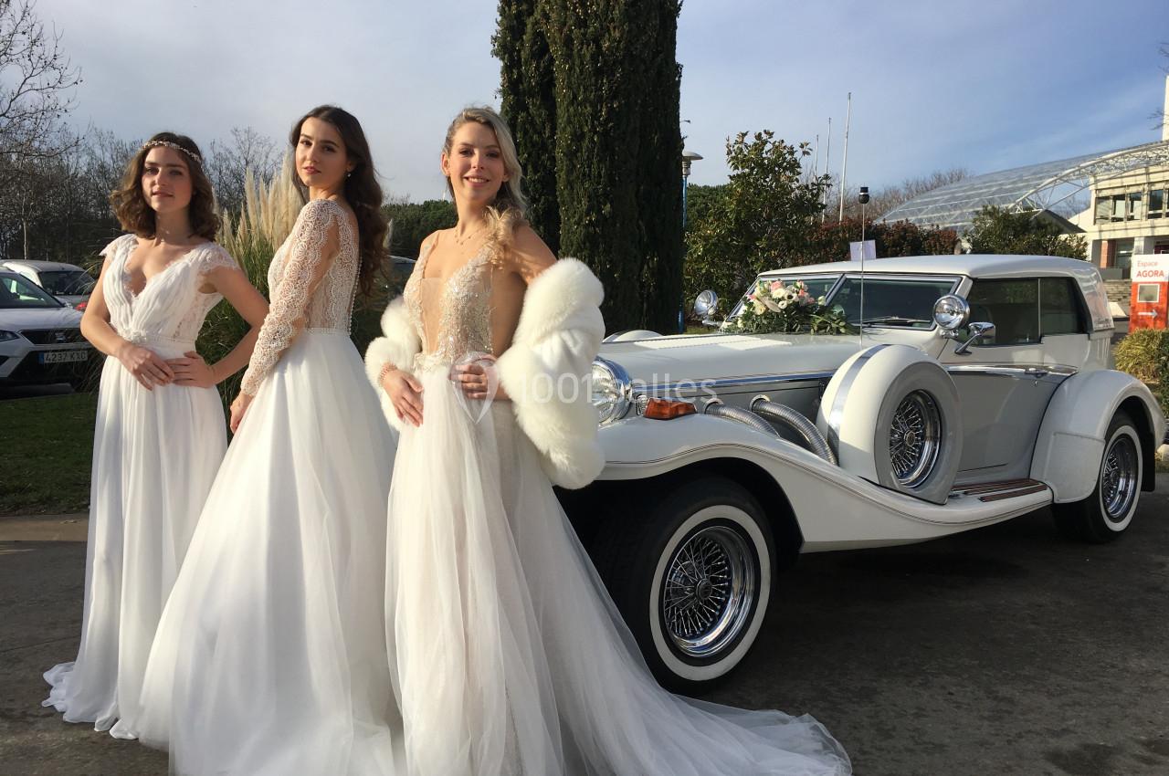 Trois femmes en robes de mariée posent devant une voiture blanche ancienne, en extérieur par temps clair.