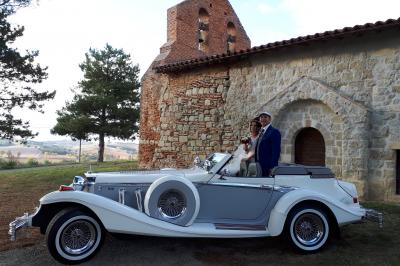Miniature Scarcityevenements #3 Un couple en tenue de mariage pose près d'une voiture ancienne blanche et grise devant une église en pierre.