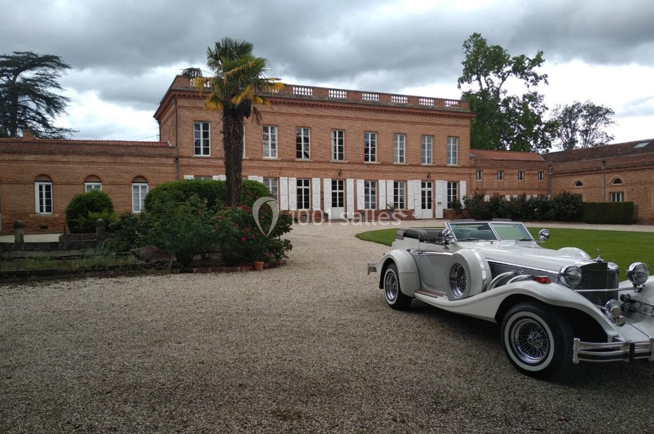 Voiture ancienne blanche garée devant un grand bâtiment en briques rouges entouré de végétation.
