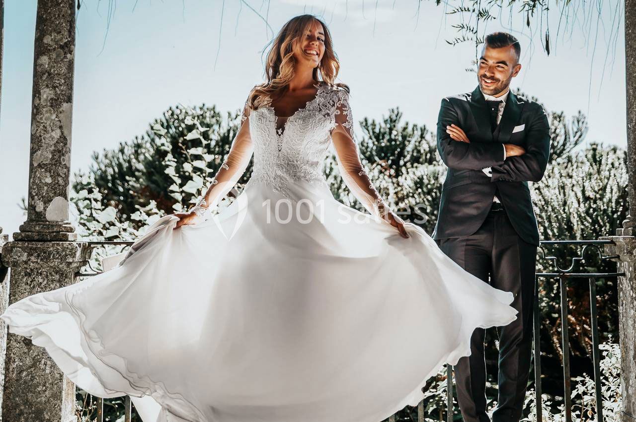 Une mariée souriante fait tournoyer sa robe blanche devant un homme en costume, en extérieur sous un ciel lumineux.