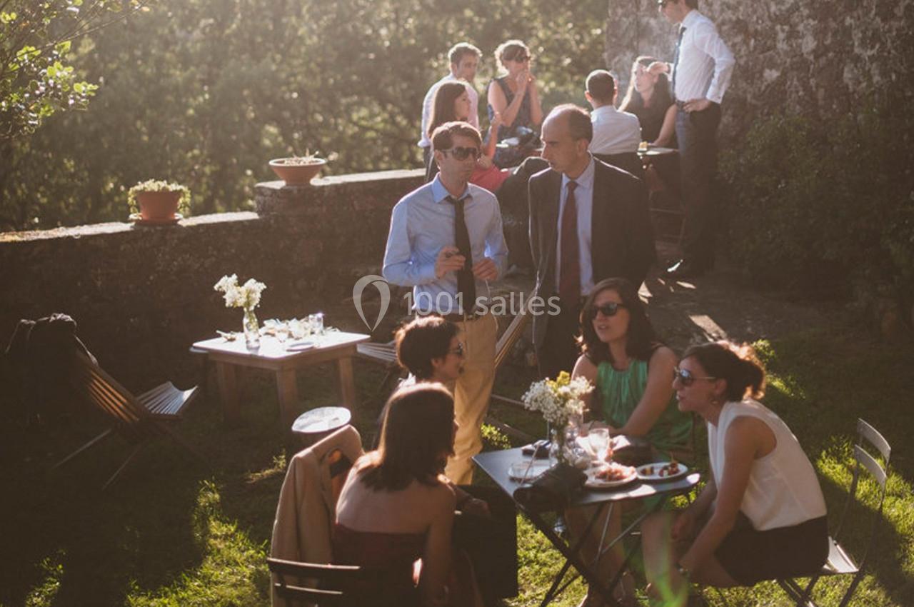 Groupe de personnes discutant autour de tables en extérieur, dans un jardin ensoleillé avec un mur en pierre.