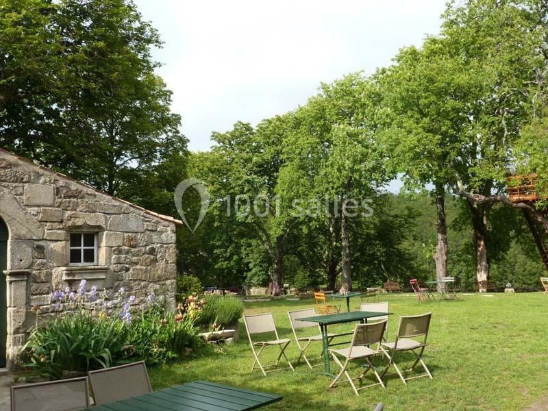 Jardin avec tables et chaises en métal, entouré d'arbres et d'une maison en pierre avec des fleurs en premier plan.