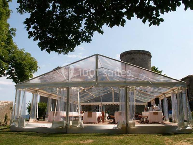 Tente transparente installée sur une pelouse, entourée d'arbres, avec des tables et des chaises à l'intérieur.