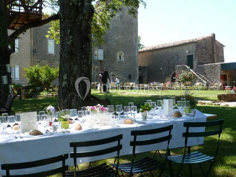 Table dressée en extérieur sous des arbres, avec chaises noires, devant un bâtiment en pierre et un jardin aménagé.