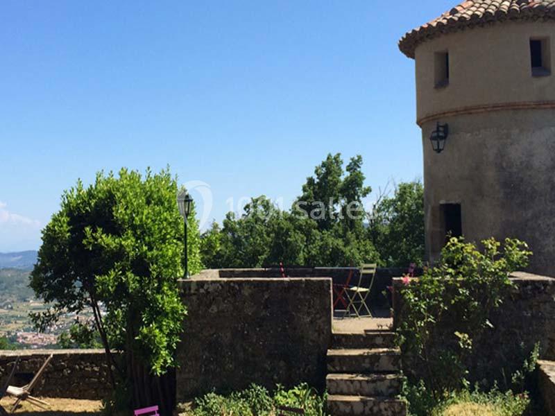 Vue d'une terrasse en pierre avec des chaises et une table, entourée de verdure et d'un bâtiment ancien sous un ciel bleu.