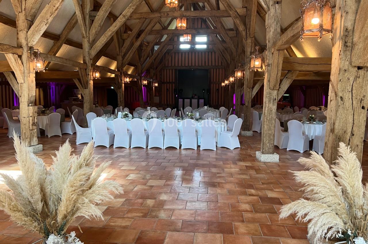 Salle de réception avec poutres apparentes, tables décorées de nappes blanches et chaises alignées, éclairage chaleureux.
