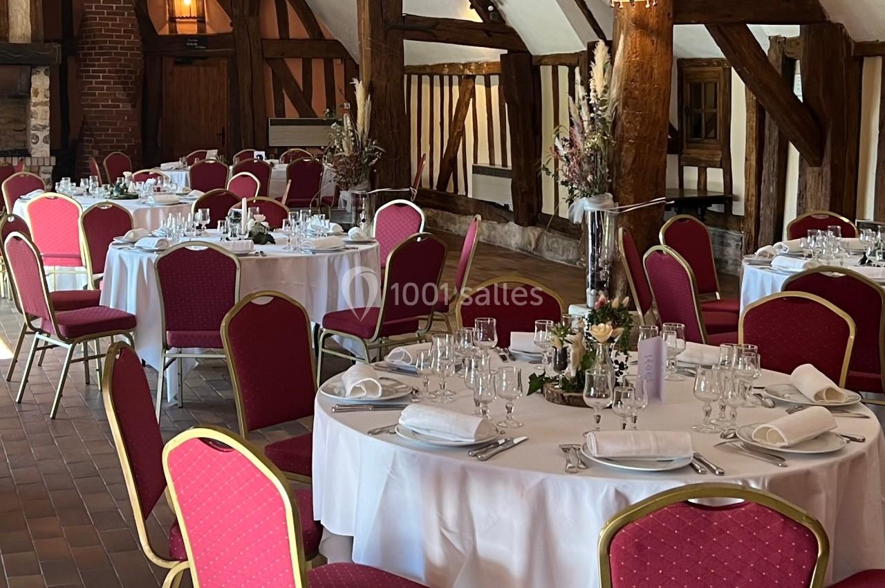 Salle de réception rustique avec tables rondes dressées, nappes blanches et chaises rouges, éclairée par des lampes murales.