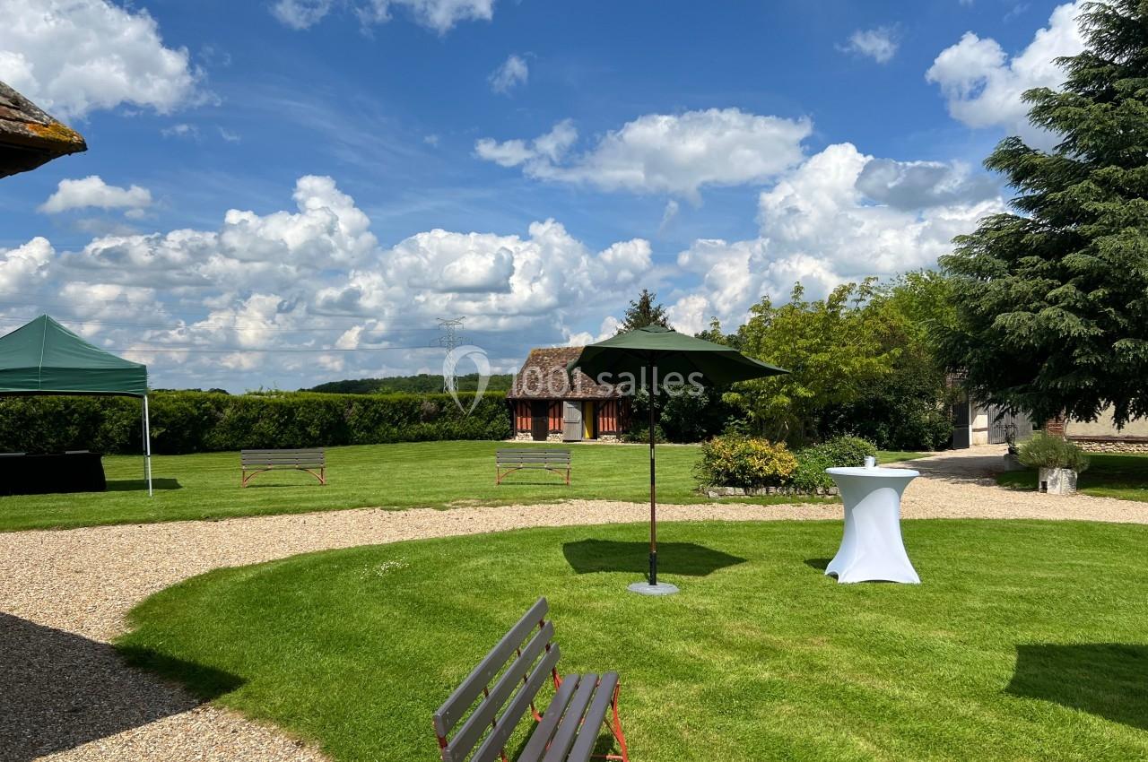Jardin verdoyant avec pelouse, bancs, parasol, table haute et ciel partiellement nuageux en arrière-plan.