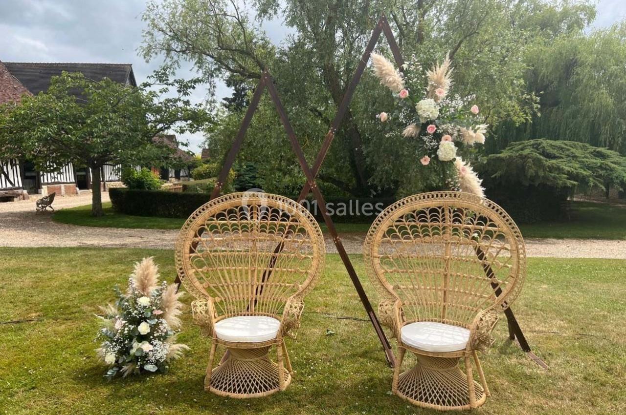 Deux fauteuils en rotin devant une arche triangulaire décorée de fleurs blanches et de pampas, dans un jardin verdoyant.