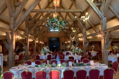 Salle de réception rustique avec poutres en bois apparentes, tables rondes dressées et chaises rouges.