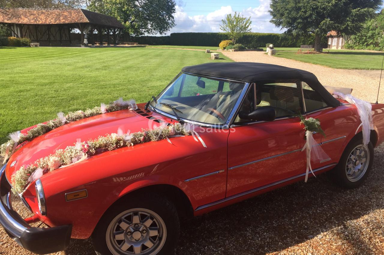 Voiture ancienne décapotable rouge décorée de fleurs blanches, stationnée sur une allée gravillonnée près d'un jardin.