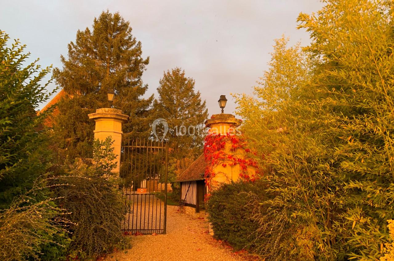 Entrée d'une propriété avec portail en fer forgé, colonnes en pierre et végétation illuminée par une lumière dorée.