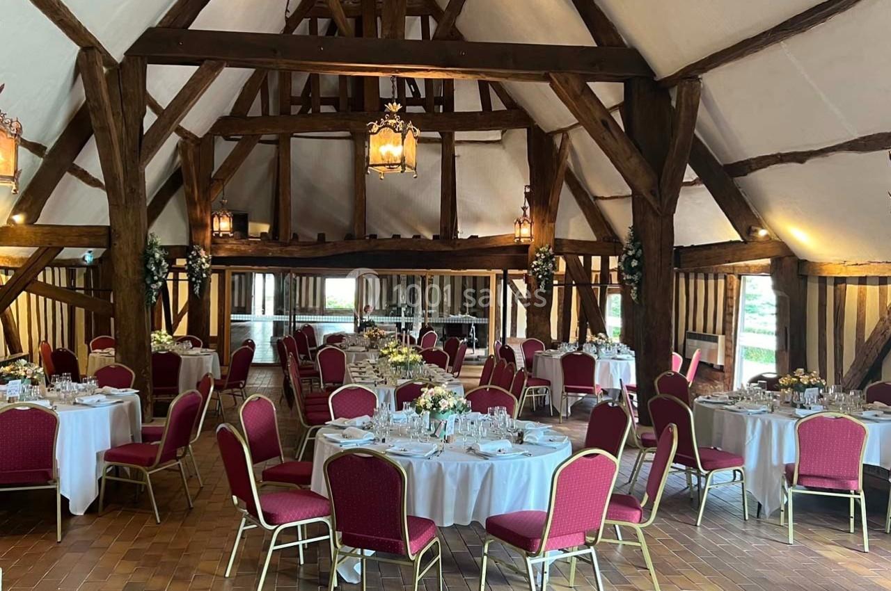 Salle de réception rustique avec poutres en bois, tables rondes dressées et chaises rouges, éclairée par des lanternes…