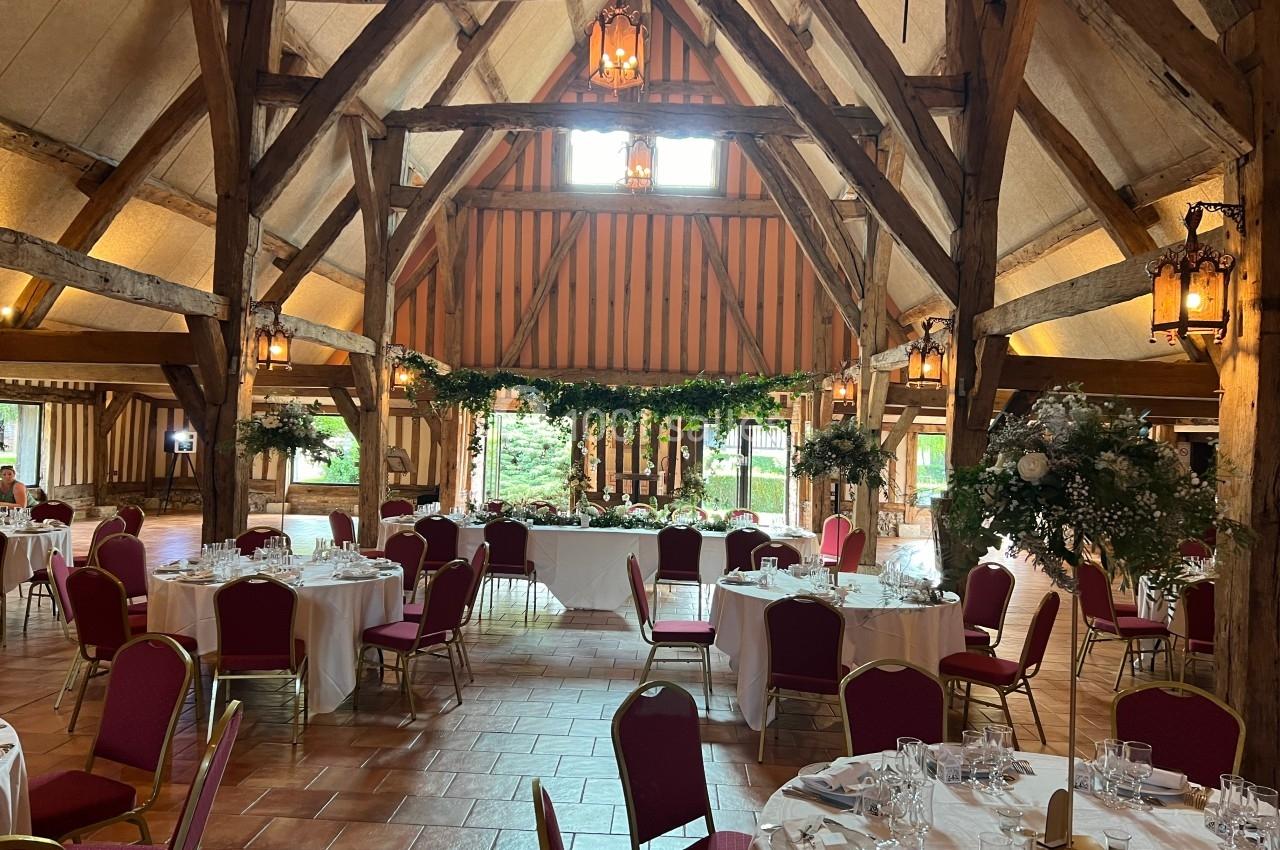 Salle de réception rustique avec poutres apparentes, tables rondes dressées et chaises rouges, décorée de fleurs.