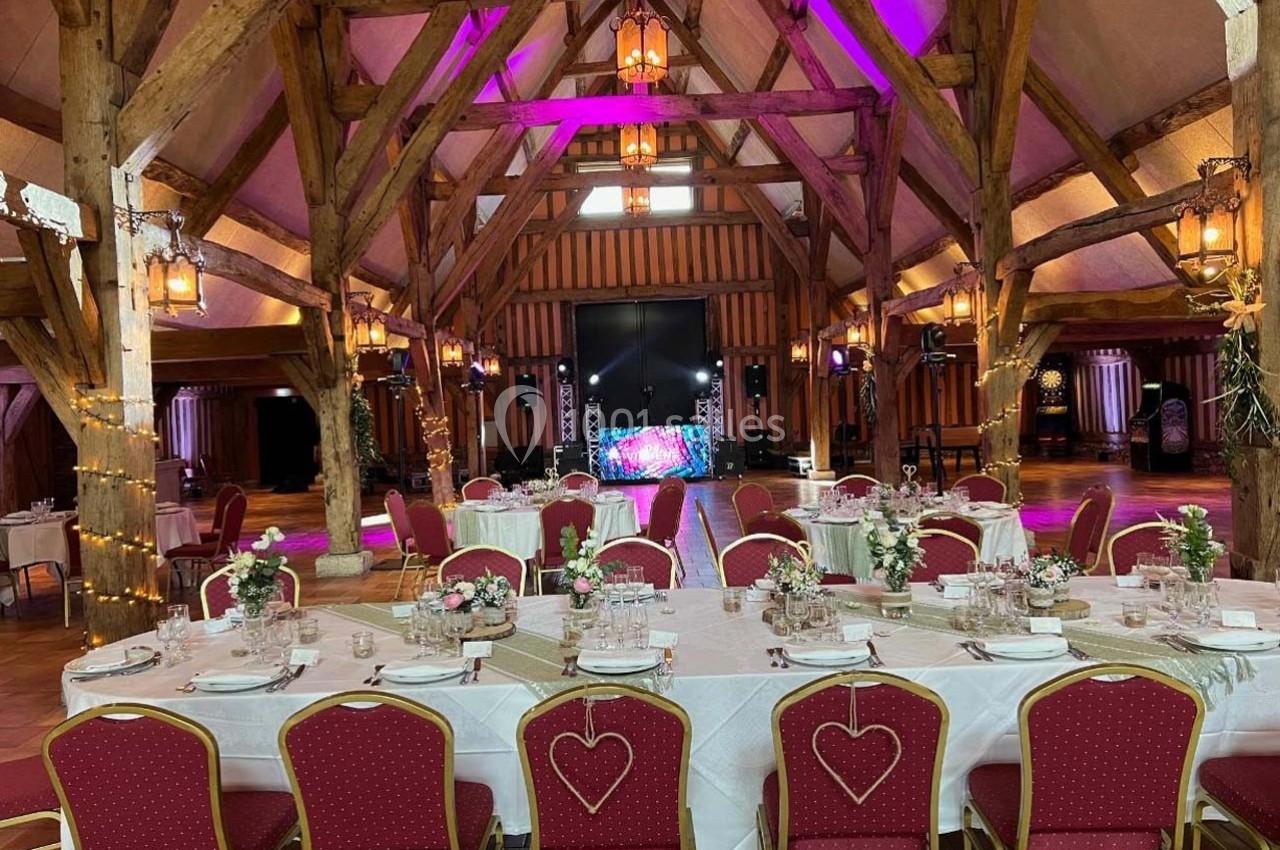 Salle de réception avec poutres apparentes, tables décorées pour un événement et éclairage coloré en arrière-plan.
