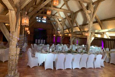 Salle de réception rustique avec poutres en bois apparentes, tables rondes dressées et chaises rouges.