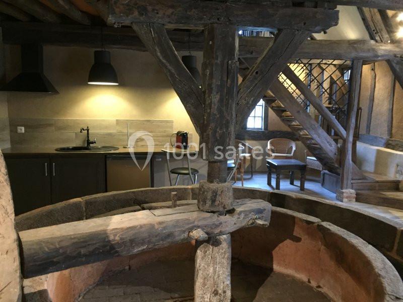 Ancien pressoir en bois dans une pièce rénovée avec cuisine moderne, table et escalier en bois visible en arrière-plan.