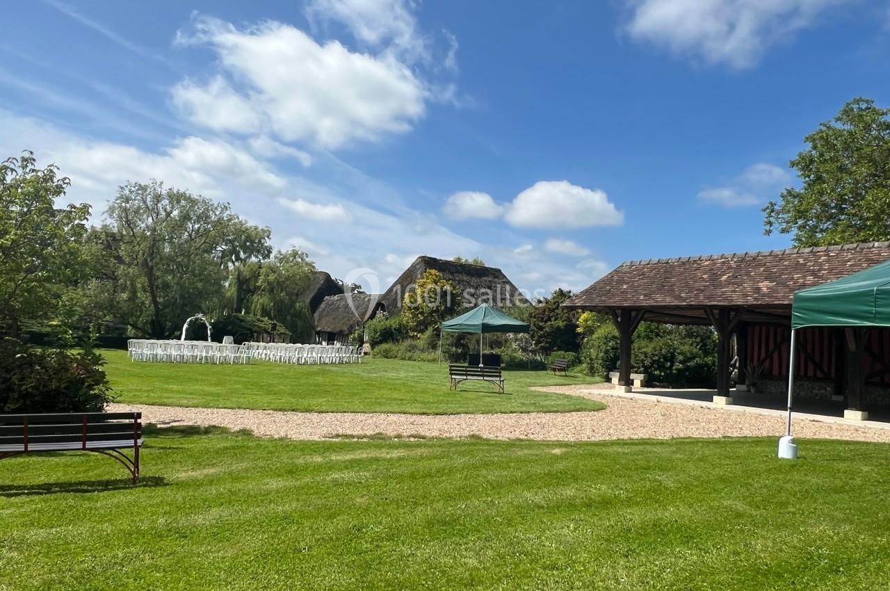 Espace extérieur avec pelouse, chaises blanches disposées pour un événement, barnums et bâtiments à toit en chaume.