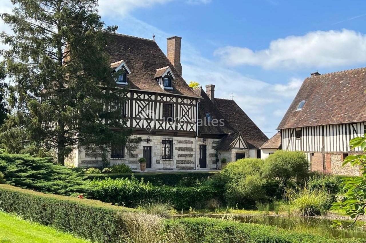 Maison à colombages entourée de verdure, avec un étang au premier plan et un ciel bleu en arrière-plan.