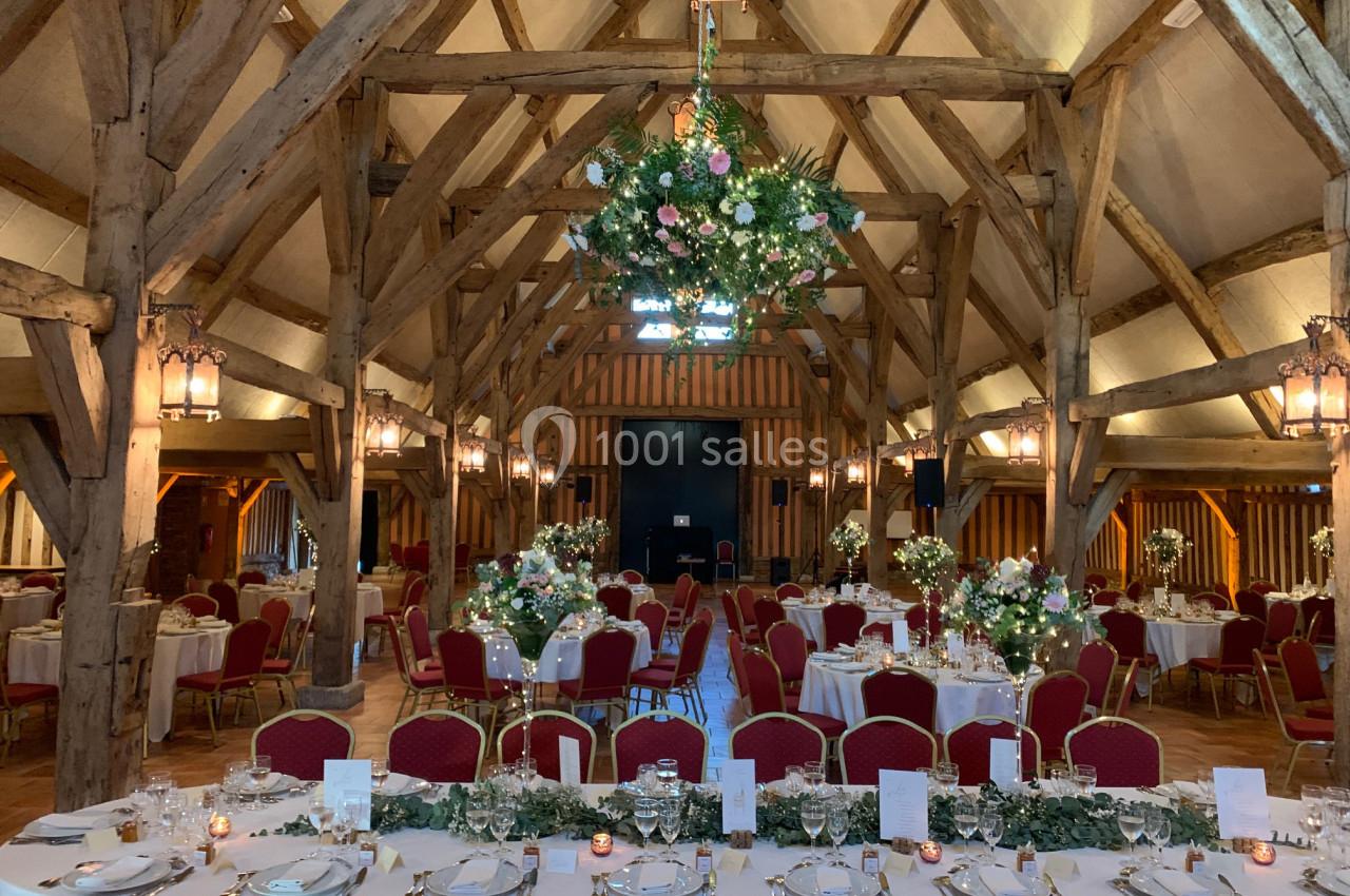 Salle de réception avec charpente en bois apparente, tables décorées de fleurs et chaises rouges.