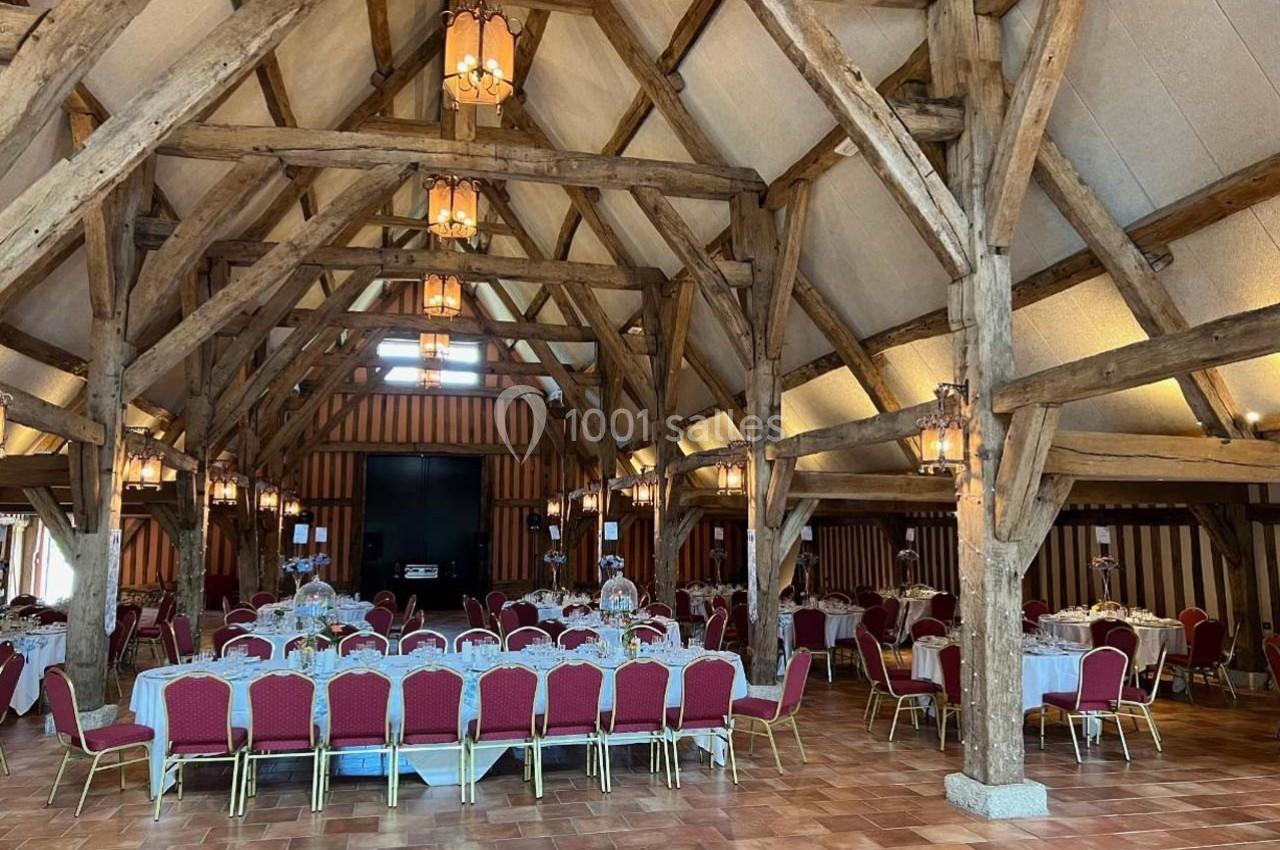 Salle de réception avec charpente en bois apparente, tables dressées et chaises rouges alignées.
