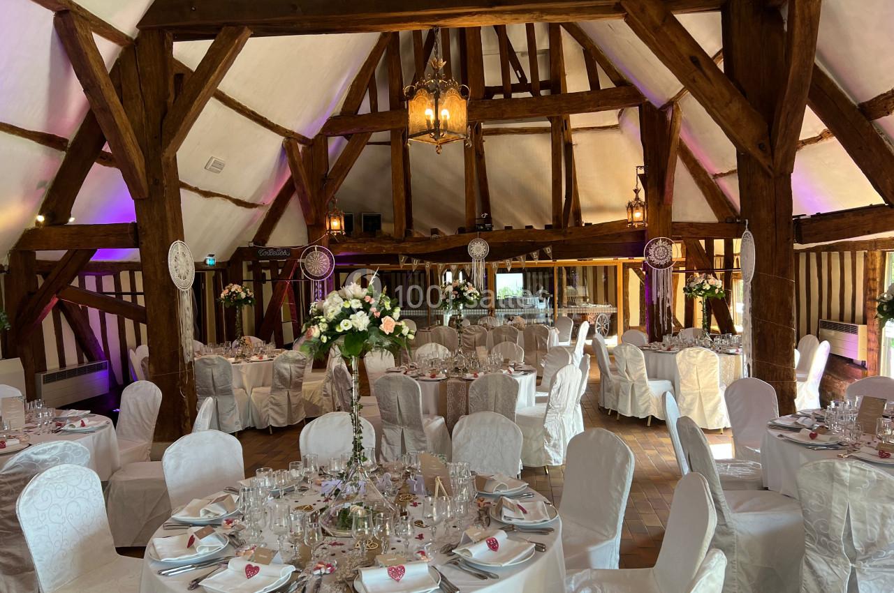 Salle de réception décorée avec des tables rondes, nappes blanches, fleurs et poutres apparentes en bois.