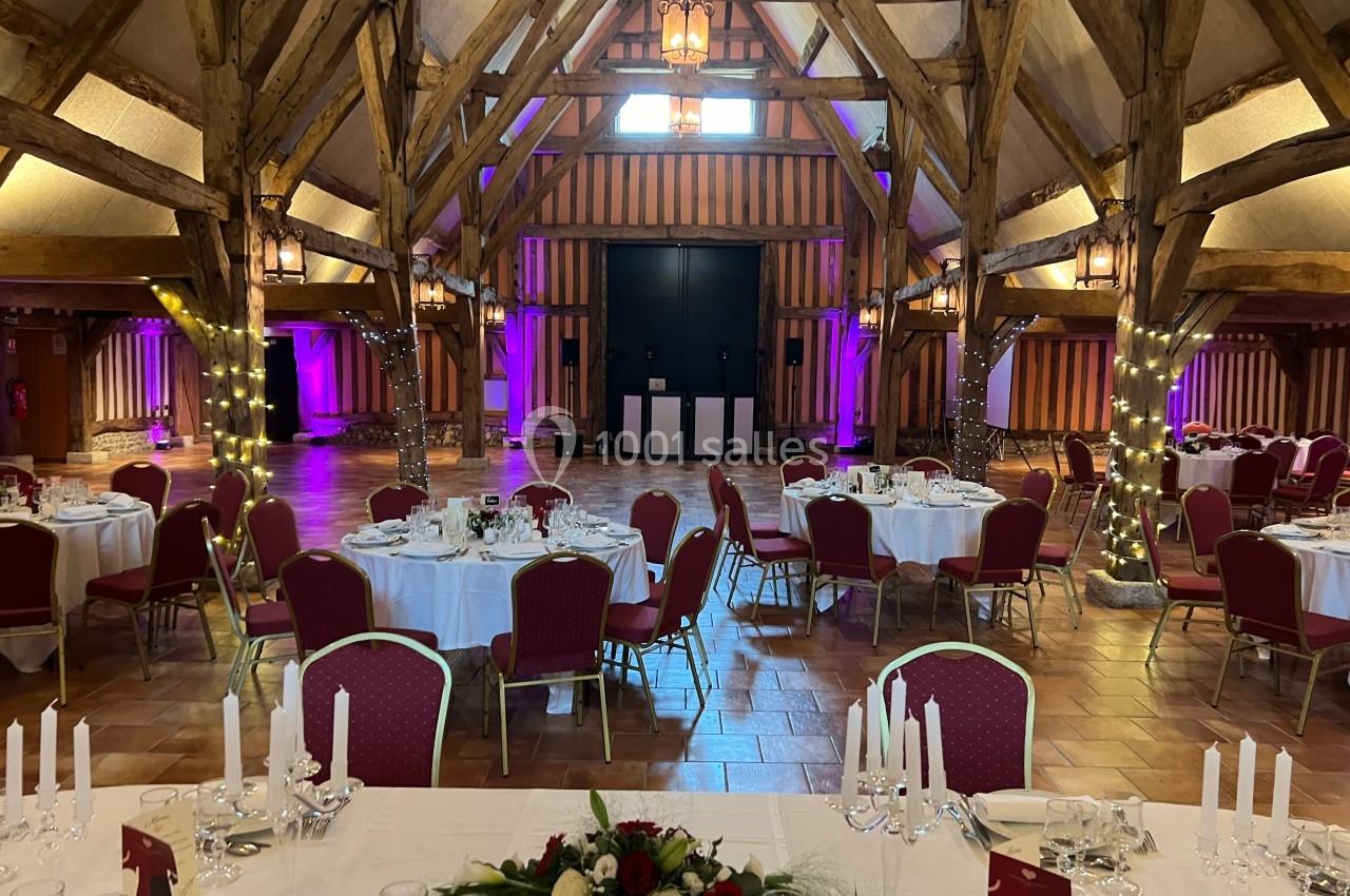 Salle de réception avec charpente en bois, tables rondes dressées et éclairages colorés sur les murs.