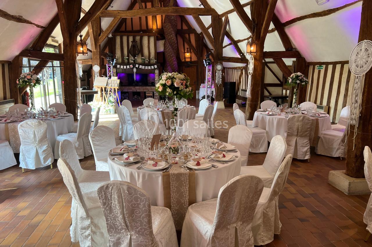 Salle de réception décorée pour un mariage avec tables rondes dressées, chaises couvertes et arrangements floraux.