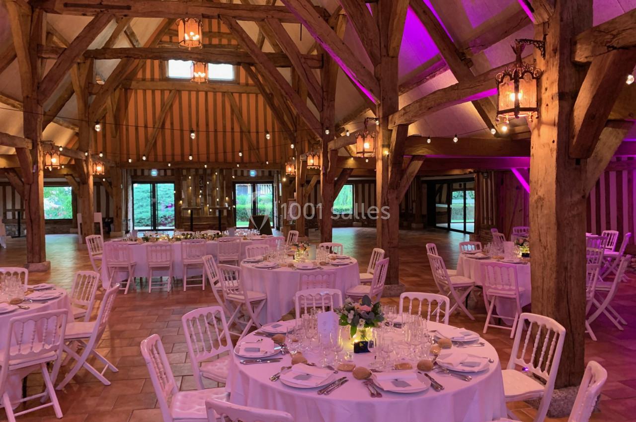 Salle de réception rustique avec charpente en bois, tables rondes dressées et éclairage tamisé violet.
