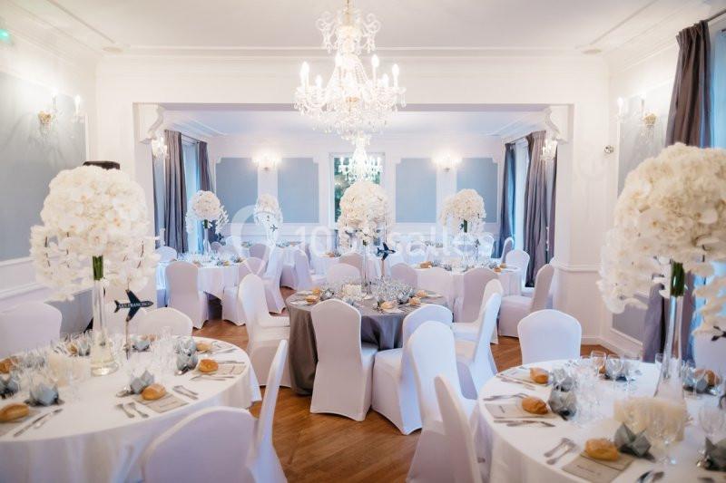 Salle de réception élégante avec tables rondes décorées de nappes blanches, centres de table floraux et chandeliers.