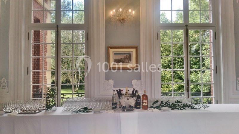 Table dressée avec des verres, bouteilles et décorations végétales devant de grandes fenêtres donnant sur un jardin.