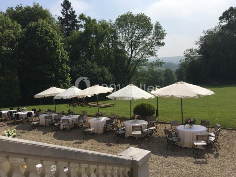 Terrasse avec tables rondes et parasols blancs, entourée de verdure et donnant sur un grand jardin arboré.