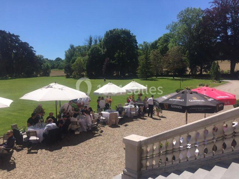 Personnes assises sous des parasols dans un jardin verdoyant par une journée ensoleillée, près d'un escalier en pierre.