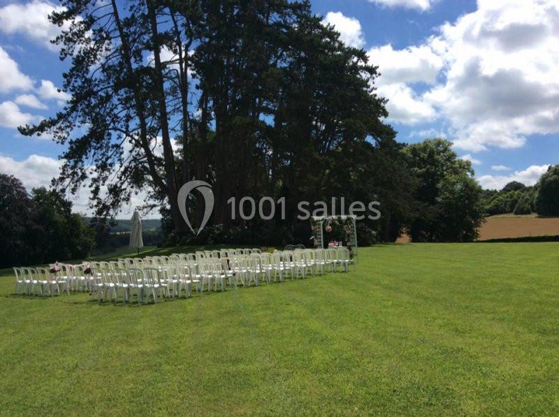 Chaises blanches disposées en rangées sur une pelouse, face à une arche décorée, sous un ciel partiellement nuageux.