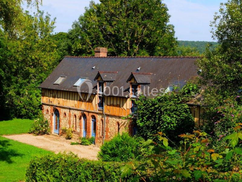 Maison en pierre et colombages entourée de verdure, avec toit en ardoise et fenêtres mansardées.