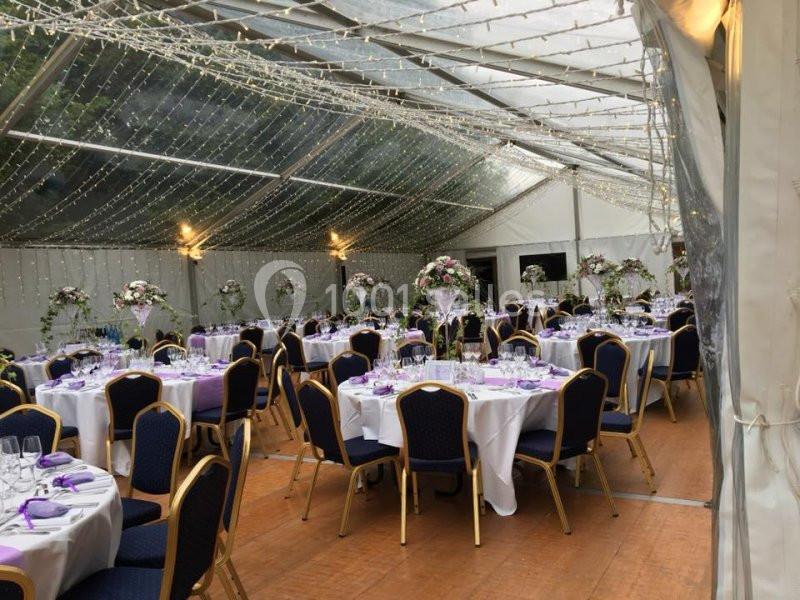 Salle de réception sous une tente transparente, décorée de guirlandes lumineuses et de tables dressées avec des fleurs.