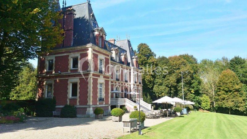 Façade d'un château en briques rouges avec un toit en ardoise, entouré de verdure et d'une terrasse aménagée.