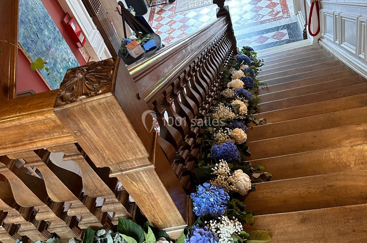 Escalier en bois décoré de fleurs blanches et bleues, menant à un hall d'entrée lumineux avec sol en carreaux.