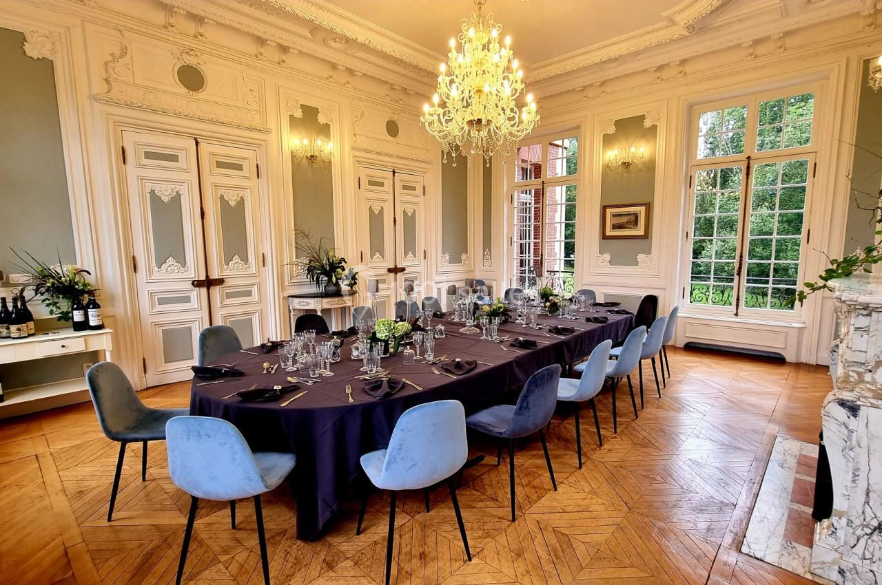 Salle élégante avec table dressée pour un repas, entourée de chaises colorées, lustre et grandes fenêtres donnant sur un…