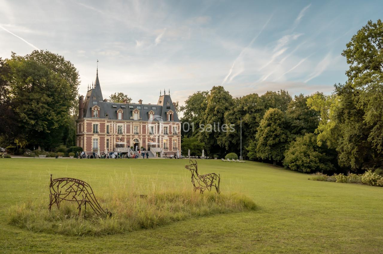 Façade d'un château entouré de pelouses et d'arbres, avec des sculptures en métal représentant des cerfs au premier plan.