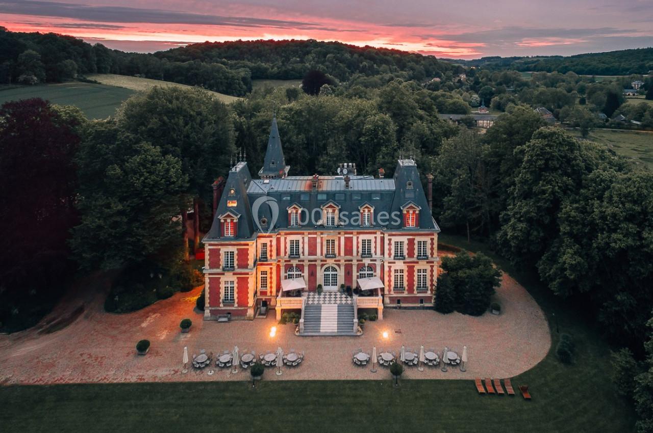 Vue aérienne d'un château entouré de verdure au coucher du soleil, avec une terrasse éclairée à l'avant.