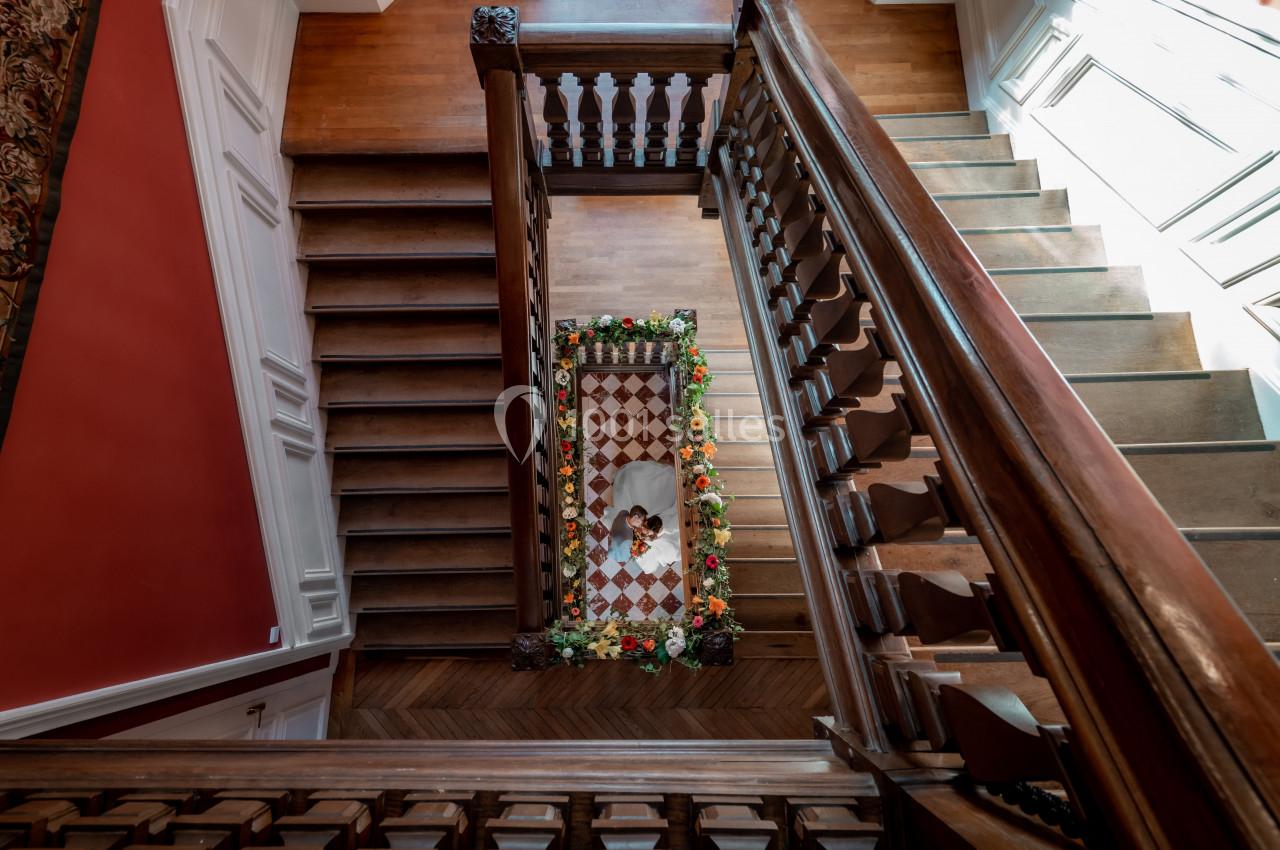 Vue plongeante sur un escalier en bois avec une rampe sculptée, menant à un sol en carreaux mosaïques entouré de fleurs.