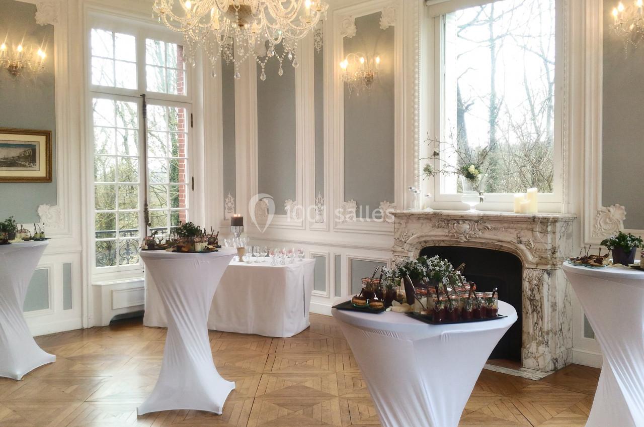 Salle élégante avec tables hautes drapées de blanc, décorée de moulures, lustre, cheminée et grandes fenêtres.