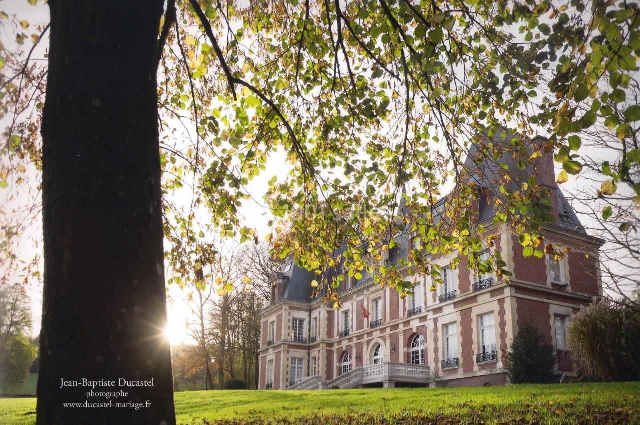 Façade d'un château en briques rouges entouré d'arbres, avec un rayon de soleil traversant les feuilles au premier plan.