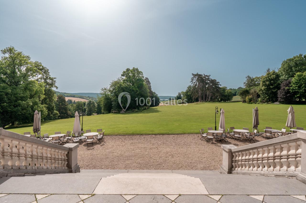 Vue depuis une terrasse sur un vaste jardin verdoyant avec des tables et parasols, entouré d'arbres sous un ciel dégagé.