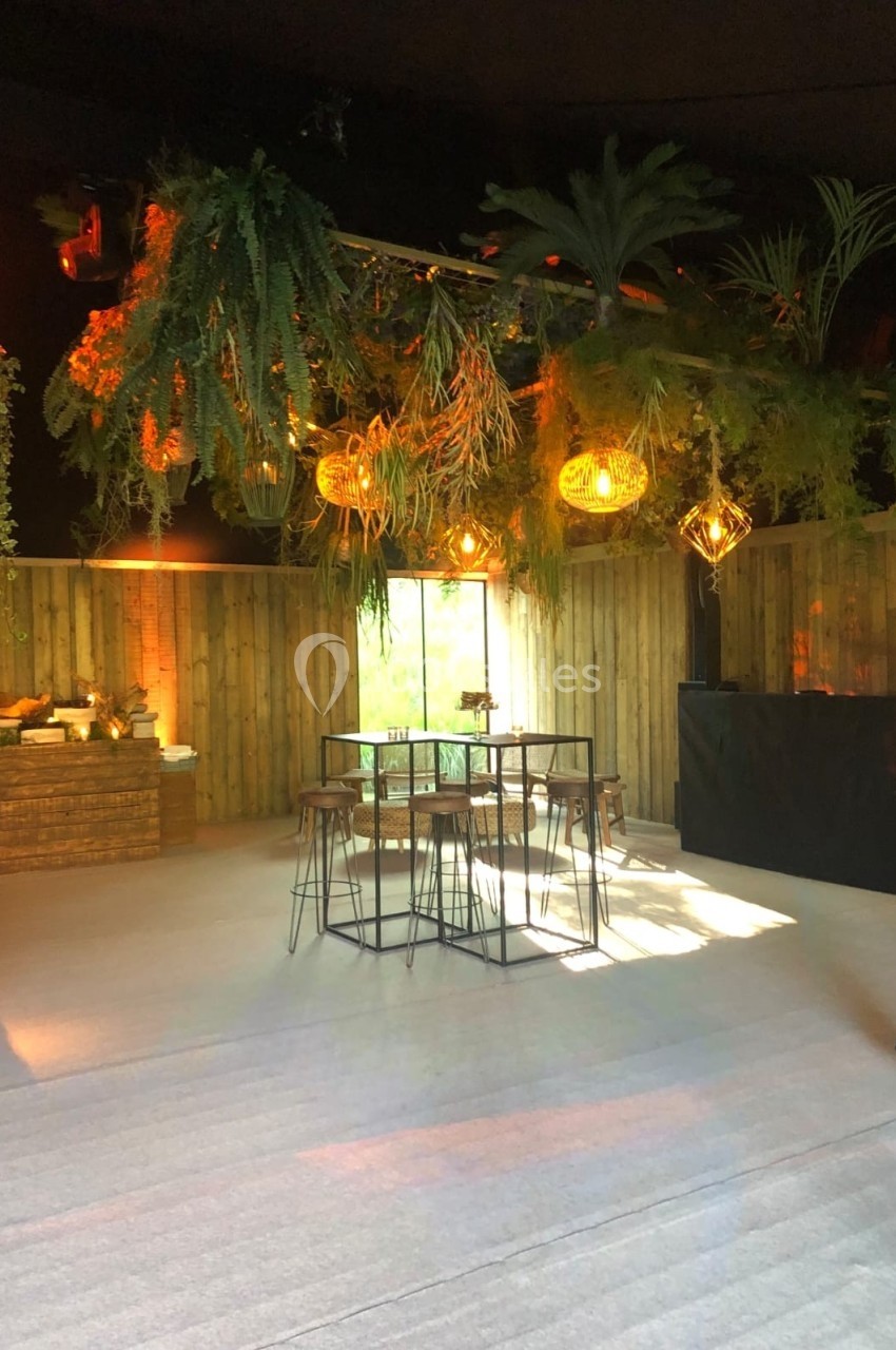 Salle décorée avec des plantes suspendues, des lampes en osier, une table haute et des chaises en métal.