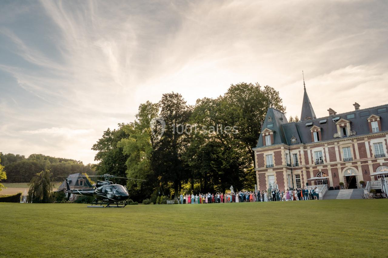 Un hélicoptère posé sur une pelouse devant un château entouré d'arbres, avec un groupe de personnes rassemblées.