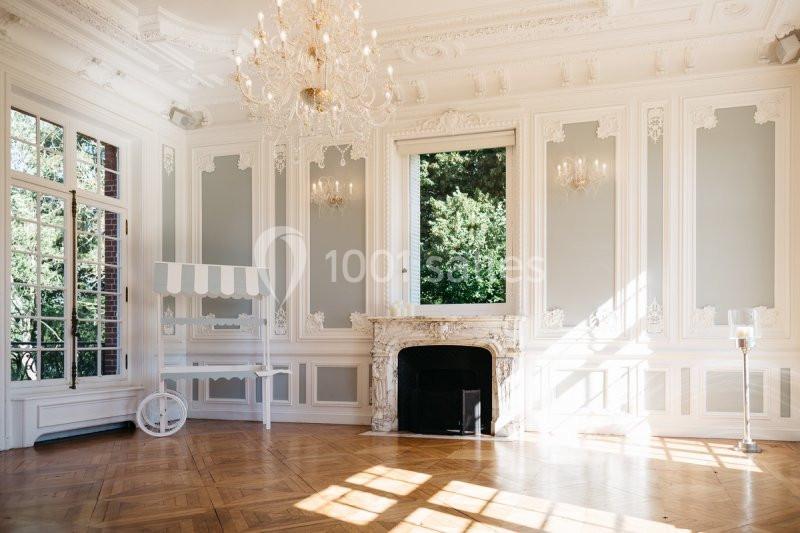 Salle lumineuse avec parquet en bois, moulures blanches, cheminée en marbre, lustre et grandes fenêtres donnant sur un…