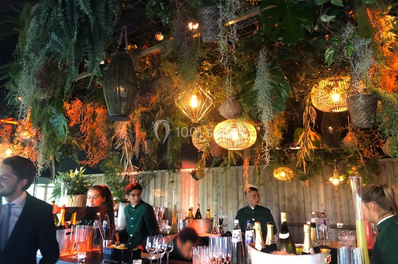 Bar décoré de plantes suspendues et lumières tamisées, avec des serveurs en uniforme et des verres disposés sur le comptoir.